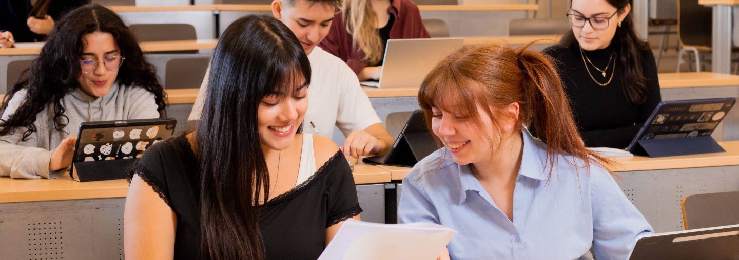 Dos estudiantes en clase sonríen mientras revisan apuntes.