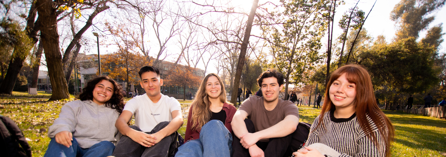 Estudiantes sonríen sentados en el pasto durante una pausa universitaria.