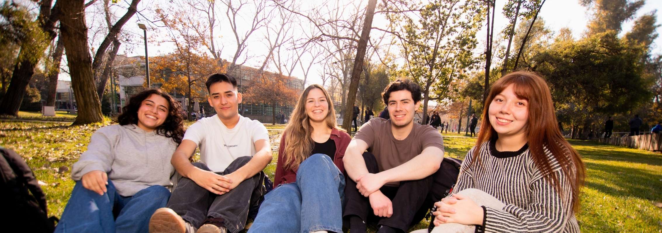Estudiantes sonríen sentados en el pasto durante una pausa universitaria.