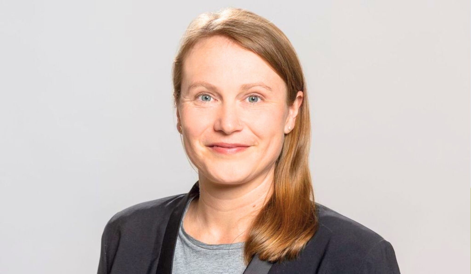 Retrato institucional de una mujer adulta, de cabello rubio y expresión amable, posando frente a un fondo gris claro